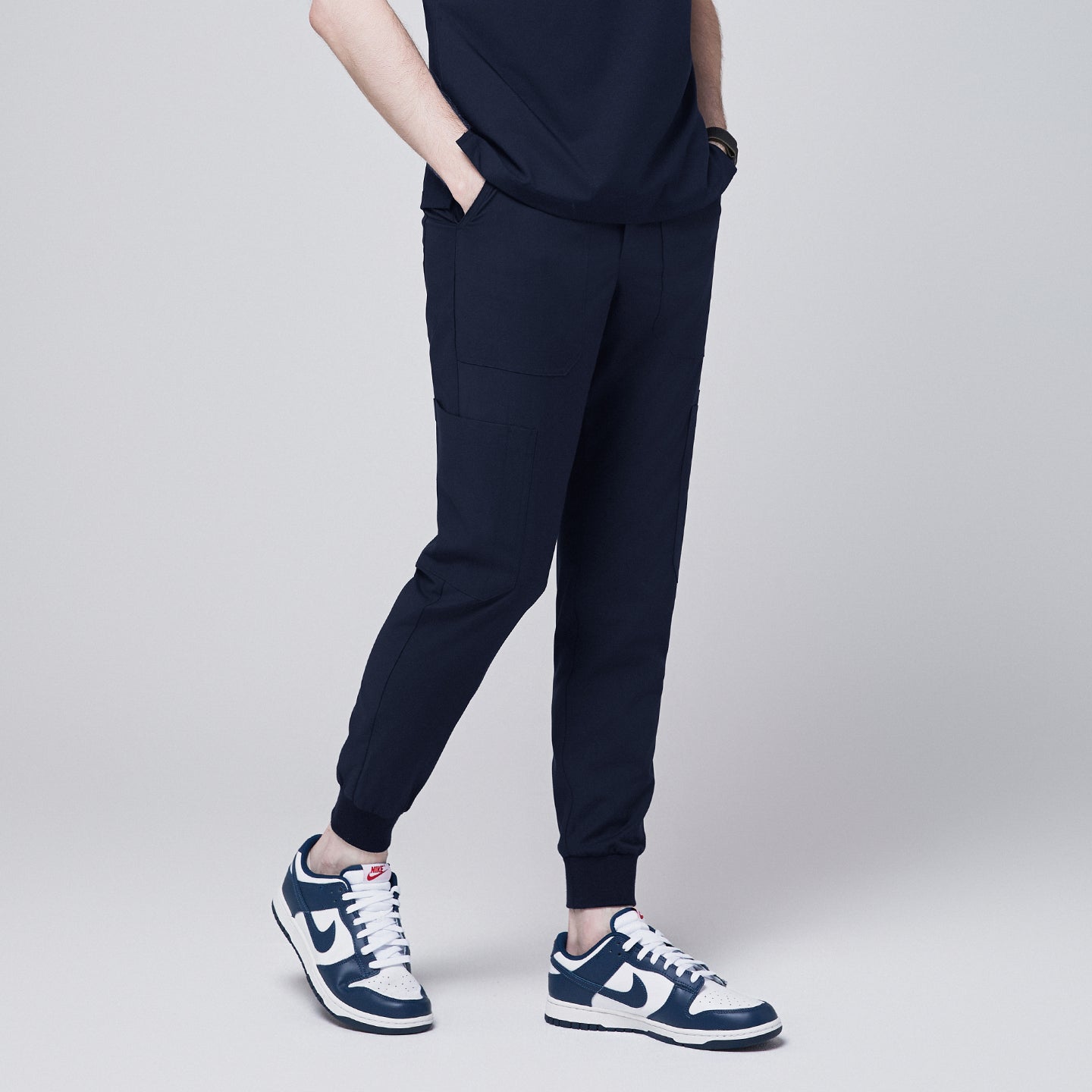 Image of Side view of navy jogger scrub pants with 8 pockets, cuffed ankles, and hands in pockets, paired with navy sneakers,Navy